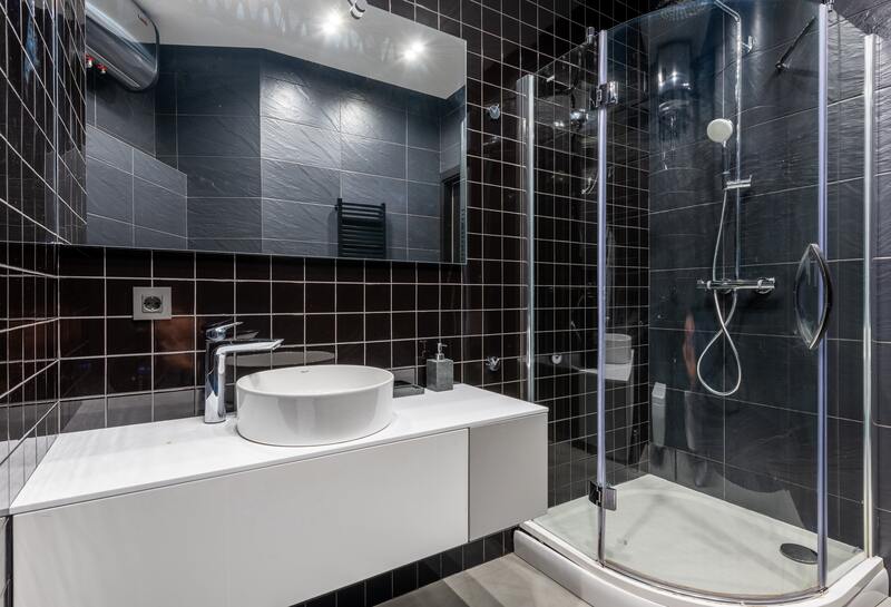 black-tiled-bathroom-round-white-sink-glass-shower-cubicle A modern black-tiled bathroom with a round white countertop basin and a corner glass shower cubicle.