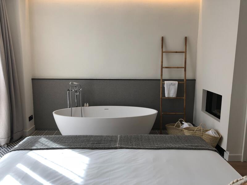 freestanding-oval-bathtub-with-wooden-ladder A freestanding oval bathtub against a grey upholstered wall with a wooden ladder towel rack and open fireplace nearby.