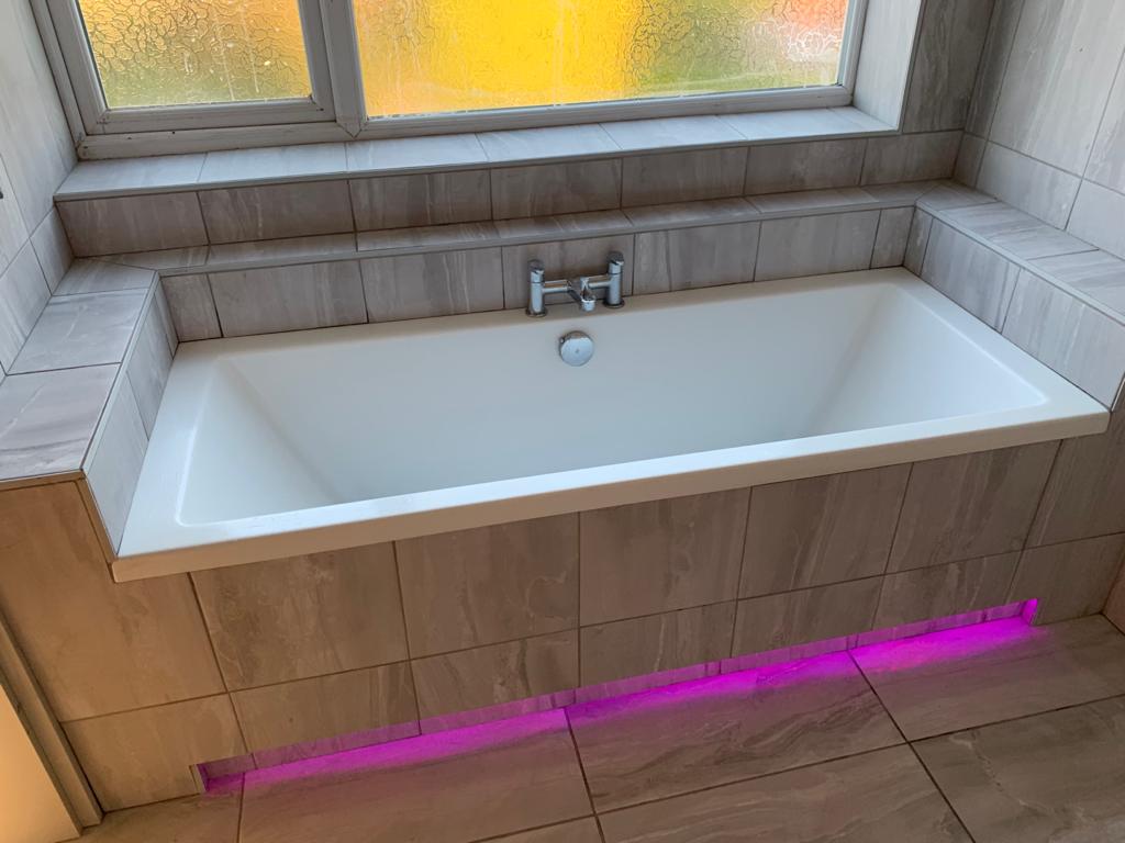 white-bathtub-tiled-surround-pink-led-strip Fitted white bathtub with grey tiled surround, chrome taps, and a pink LED light strip along the floor edge.